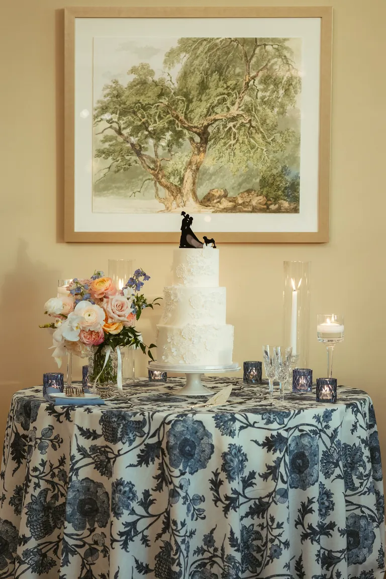A three-tier wedding cake on a blue floral tablecloth beneath a painting
