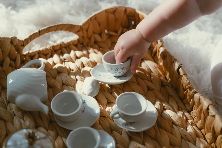 Small hands pouring from a toy tea set on a woven tray