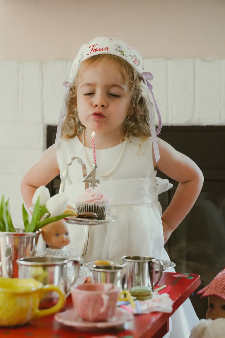 A girl in a lace crown blowing out a candle at her birthday tea party