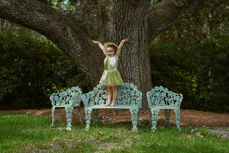 A girl in a party dress standing with her arms out on a garden bench beneath a live oak