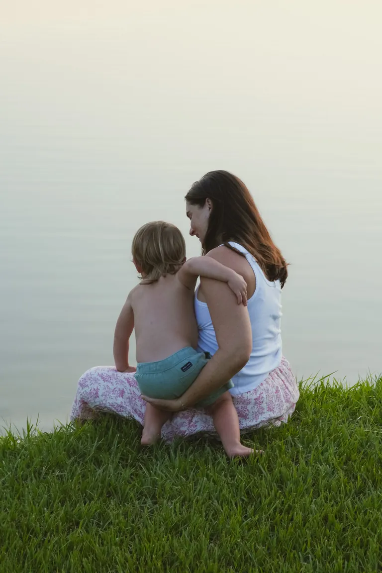 A mother and her child sitting together in the grass by the water
