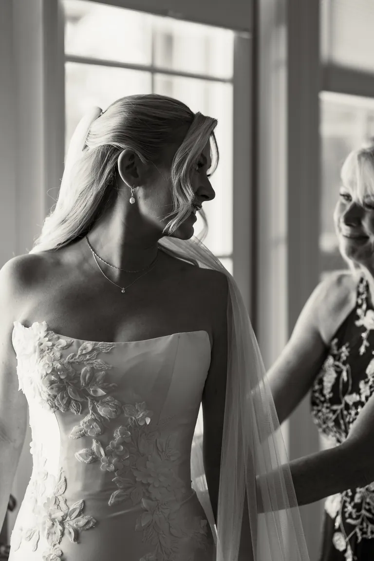 A bride in a beaded gown being helped into it by the window, black and white