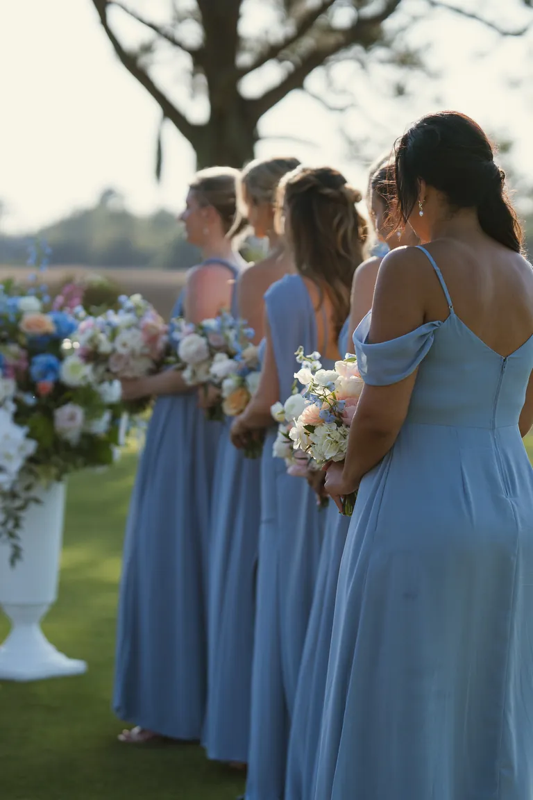 Bridesmaids in dusty blue dresses holding pastel bouquets under the oaks